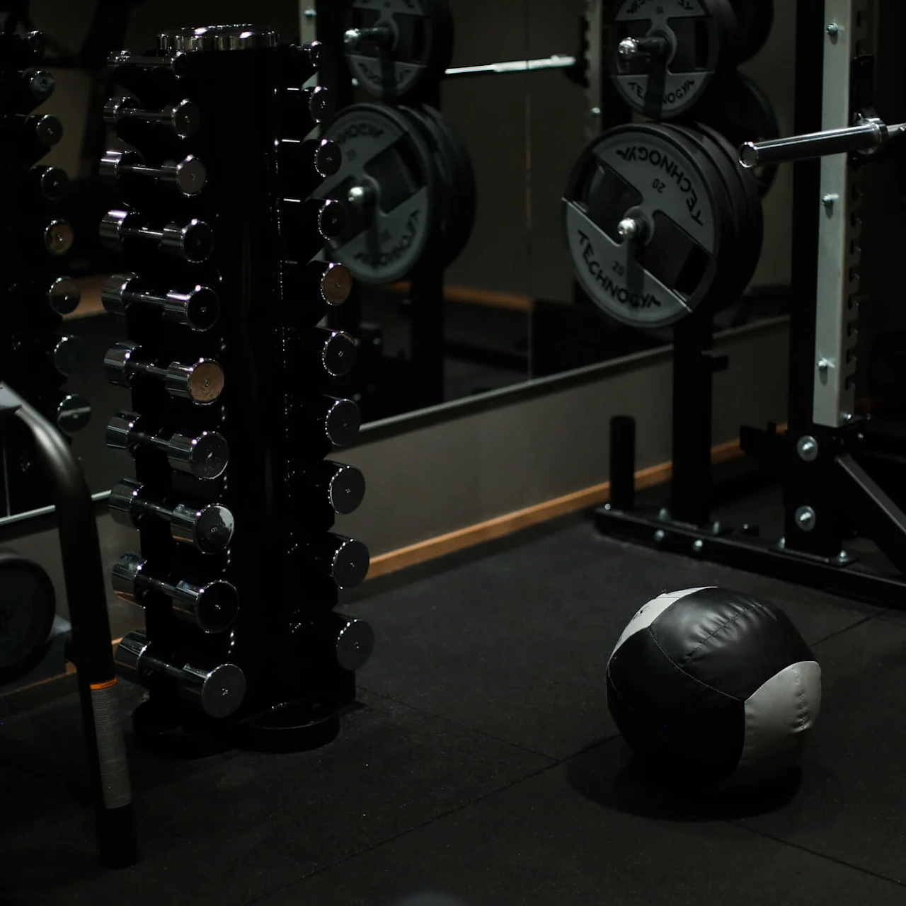 A rack of dumbbells stands in a dimly lit gym, reflecting in mirrors. Nearby, a barbell with weight plates and a medicine ball rest on the rubber floor.