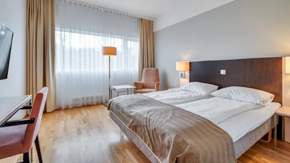 A tidy hotel room with a large bed, armchair, and a table under ambient lighting near a curtained window.