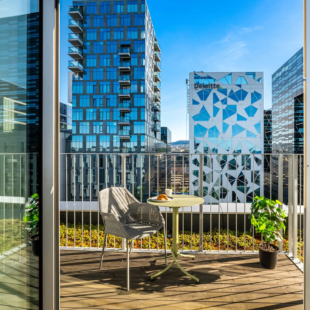 A small balcony with a table, a croissant, and coffee. Surrounded by modern high-rise buildings under a clear blue sky. Visible text: "Deloitte".