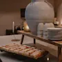 Ett stort keramikkärl står på ett träställ bredvid staplade koppar och tallrikar. Nedanför fyller en bricka med bakverk bänkskivan. Tända ljus skapar en varm, inbjudande stämning.