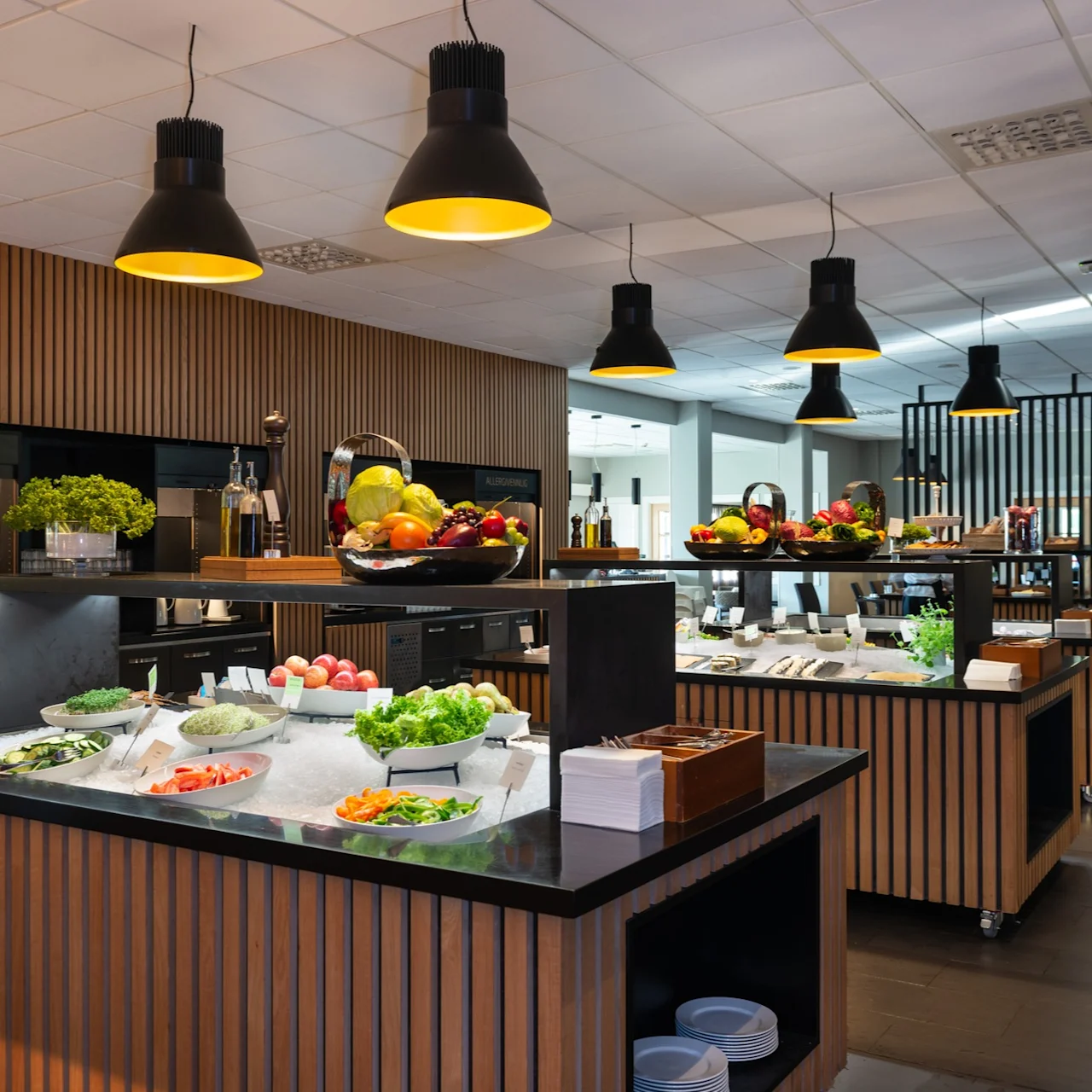 Buffet stations display assorted fresh fruits, vegetables, and salads on ice trays; modern dining area with wooden accents and overhead black pendant lights.