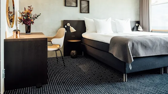 Et moderne hotelværelse har en pænt redt seng med hvide puder, ledsaget af et skrivebord med en vase med farverige blomster. Grå toner dominerer den rolige og elegante indstilling.