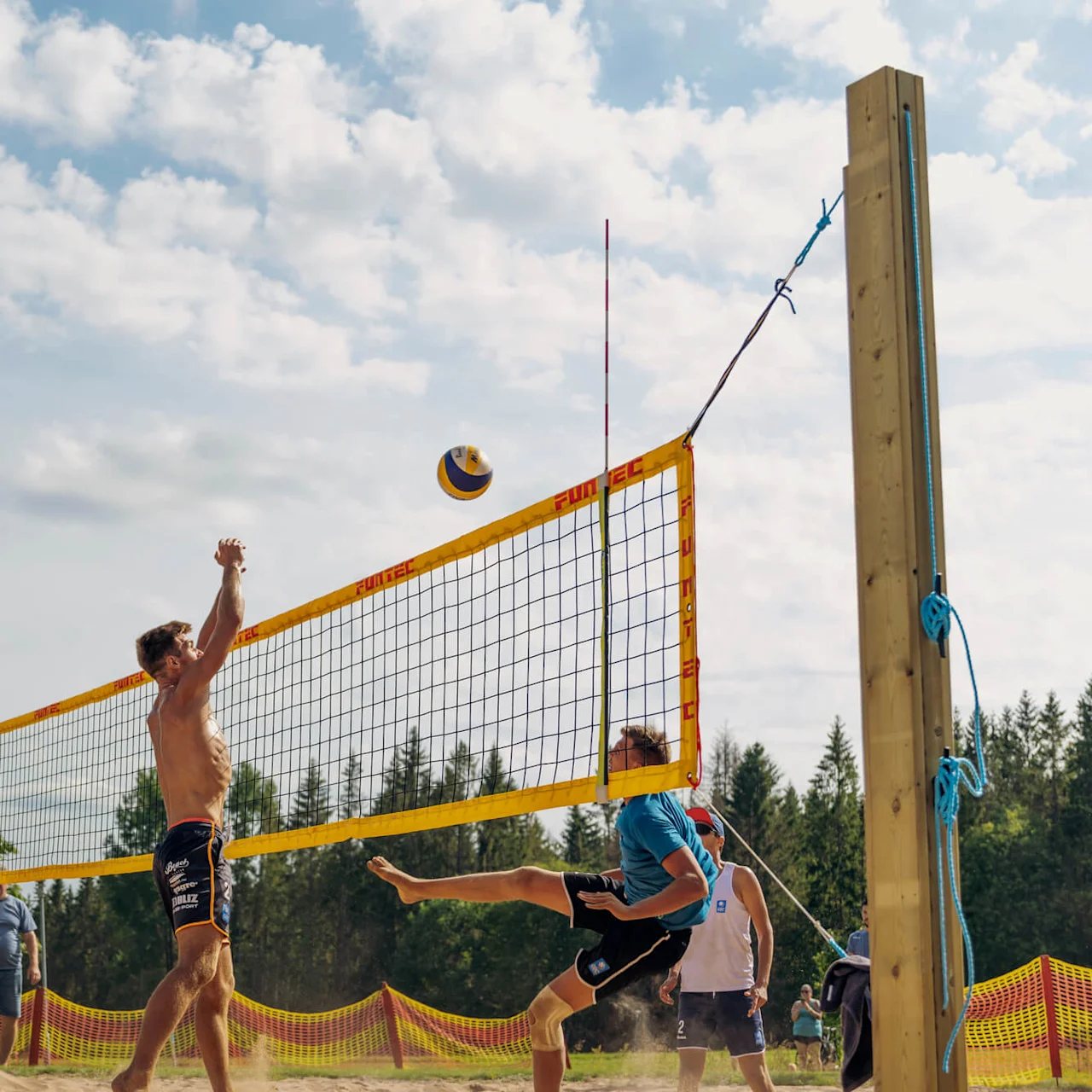 En skjorteløs mann hopper for å blokkere en volleyball på en sanddekket utendørsbane. En dommer og trær er synlige i den solfylte bakgrunnen. "SWE VOLLEYBALL" og "FUNTEC" står på nettet.