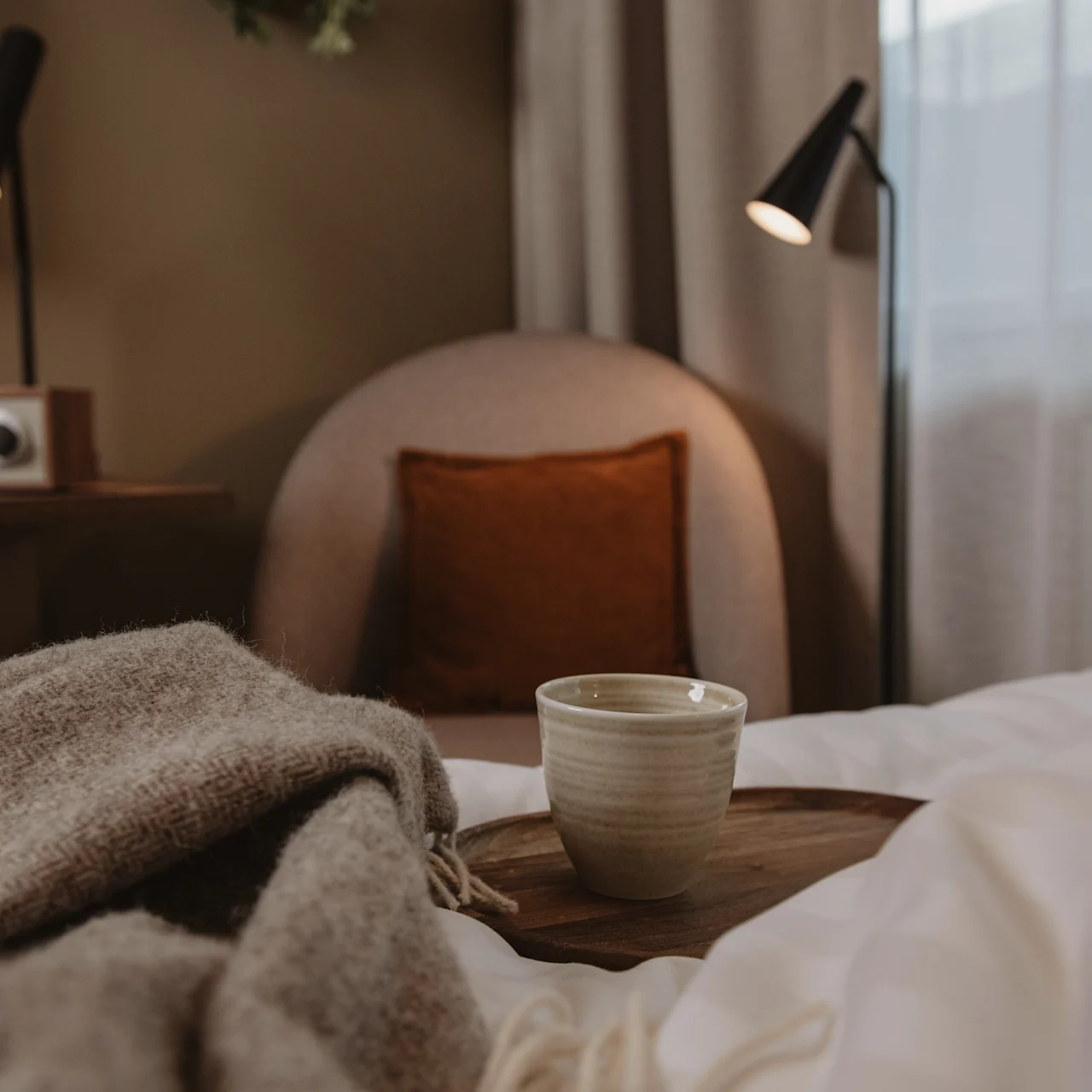 A ceramic mug sits on a wooden tray on a bed with a textured blanket, surrounded by a cozy chair, lamps, and a radio in a softly lit bedroom.
