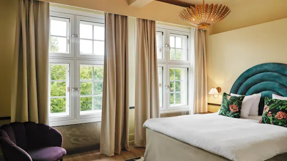 Bed stands neatly in a bright room with large windows and beige curtains, under a stylish golden chandelier, complemented by a teal headboard and floral cushions.