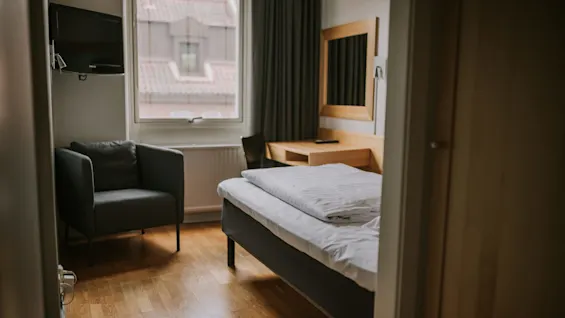 En pænt redt seng med hvide lagner er i et hotelværelse; i nærheden står en grå lænestol og et skrivebord med en stol under et vindue.