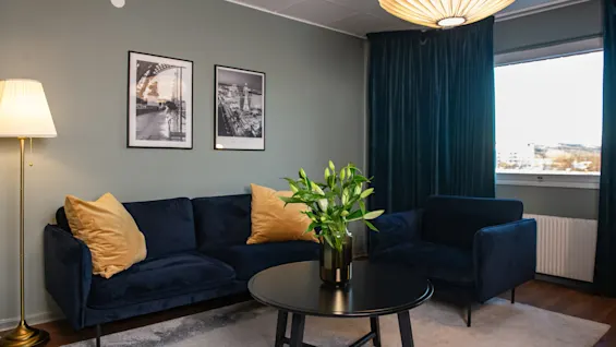 A dark blue sofa and chair sit beside a black coffee table with a vase of green plants. Wall art includes photos labeled “LAS VEGAS”. The room has dark curtains and warm lighting.