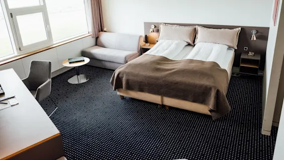 En pænt redt dobbeltseng med brunt sengetøj står under et vindue, ledsaget af en grå sofa og rundt bord, på et mørkt gulvtæppe.