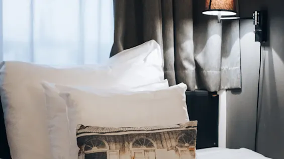 A neatly made bed with white pillows and a decorative pillow featuring a door print, illuminated by a wall lamp, set against a backdrop of gray curtains and soft natural light.