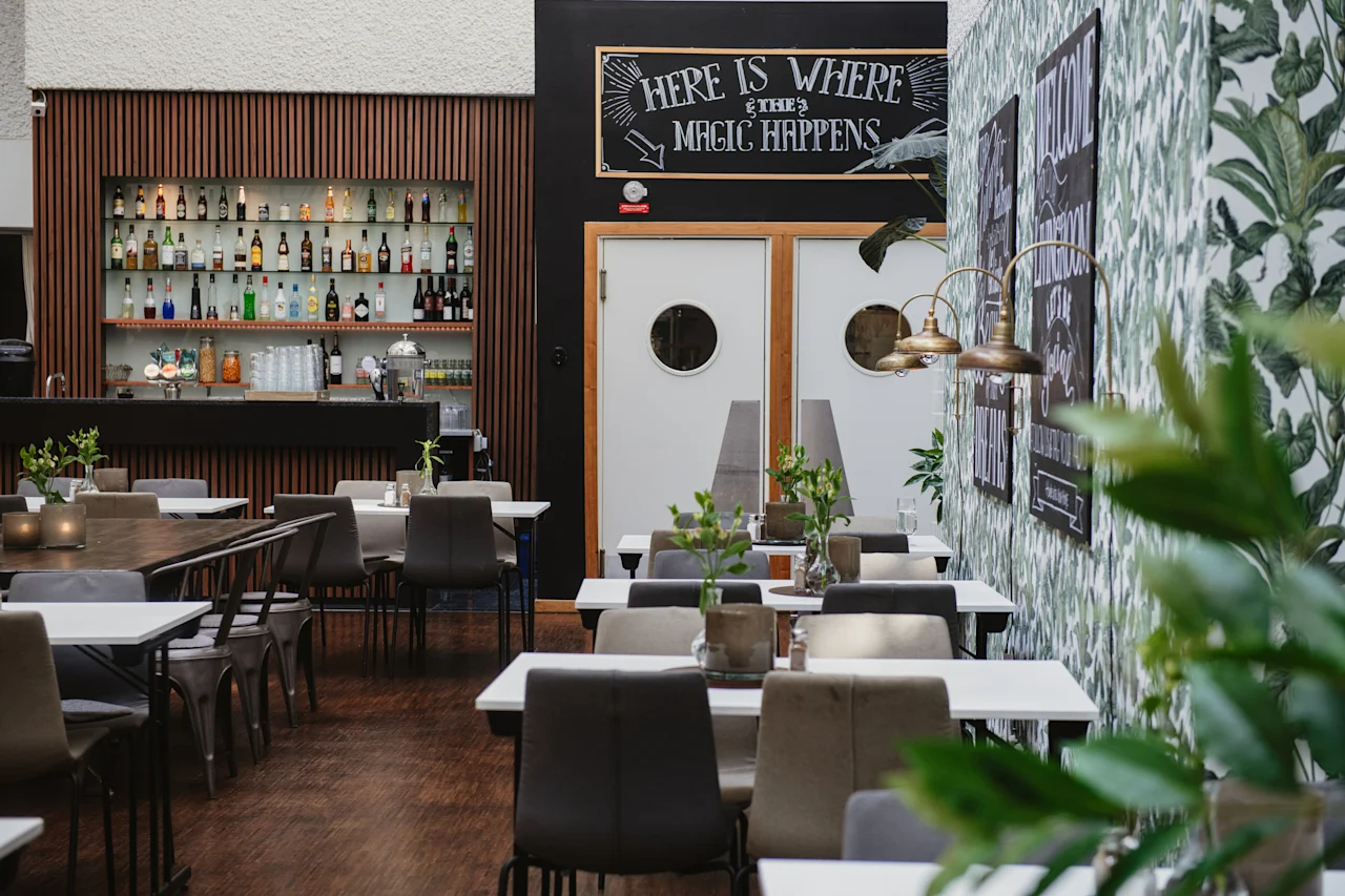En modern restaurang med prydligt arrangerade bord och stolar. Hyllor med färgglada spritflaskor pryder baren. Text på en griffeltavla lyder: "HERE IS WHERE THE MAGIC HAPPENS."