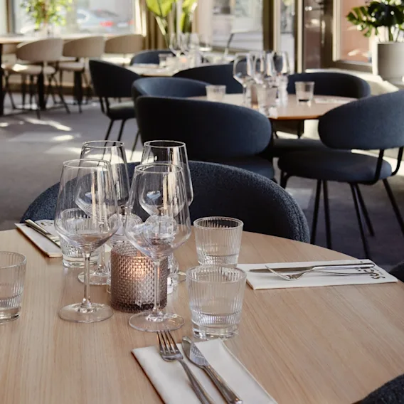 En rund trätabell är uppsatt med vinglas och bestick på servetter märkta "SOCIAL." Stolar omger bordet i en ljus, växtfylld matsal.