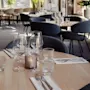 A round wooden table is set with wine glasses and cutlery on napkins labeled "SOCIAL." Chairs surround the table in a bright, plant-filled dining area.