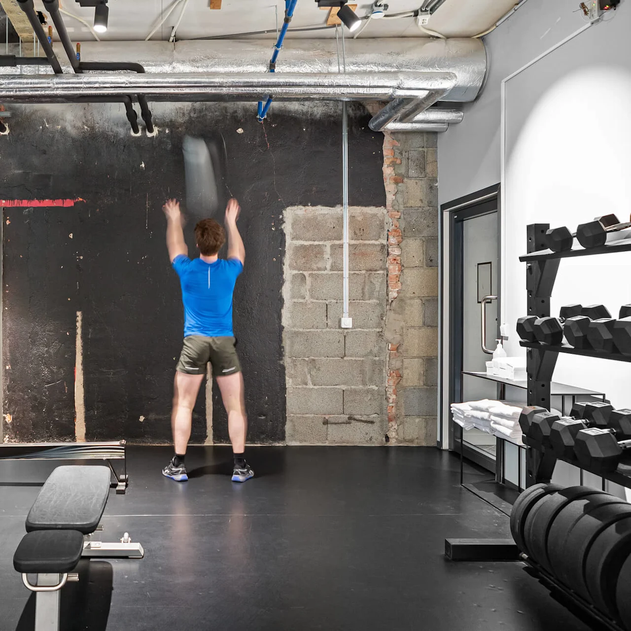 En person i blå tröja tränar genom att kasta en boll mot en svart vägg i ett gym med bänkar, vikter och industriella rör.