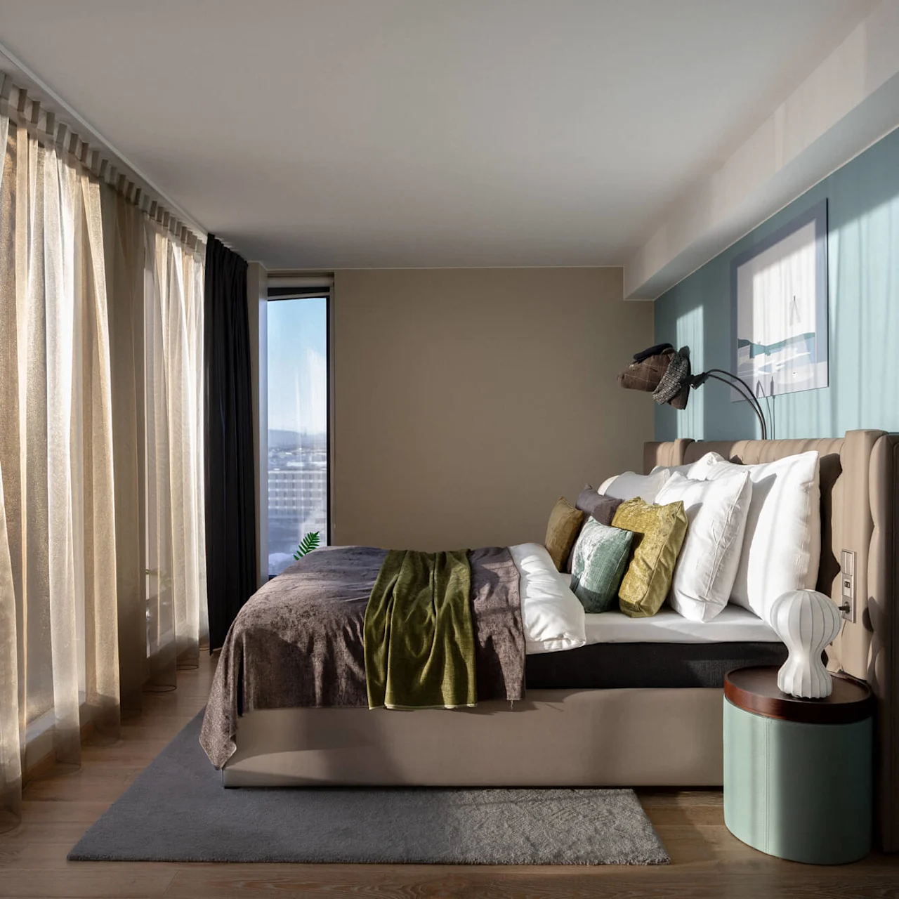 A neatly made bed with colorful pillows and a blanket sits beside a small lamp on a round table. Soft light filters through sheer curtains in a modern bedroom.