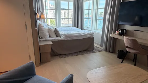 A bright hotel room with a bed by a large window overlooking a body of water and city buildings. An armchair and coffee table are in the foreground.