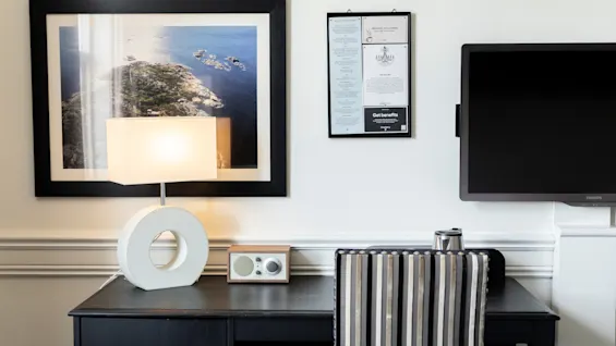Et hotelværelses skrivebordsområde med en lampe, radio og et indrammet kystlandskabsbillede. En vægskærm viser Få fordele.