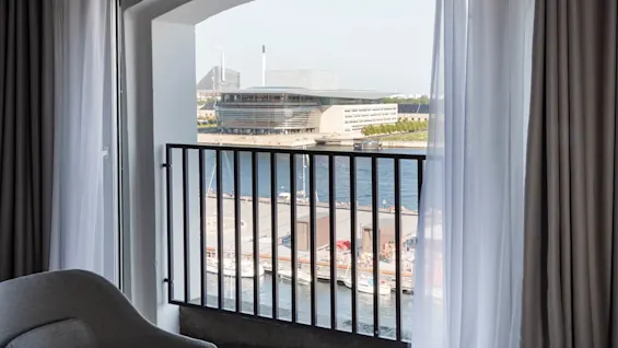 A balcony window, overlooking a harbor, frames a modern architectural building with boats moored by a dock under a clear sky. 