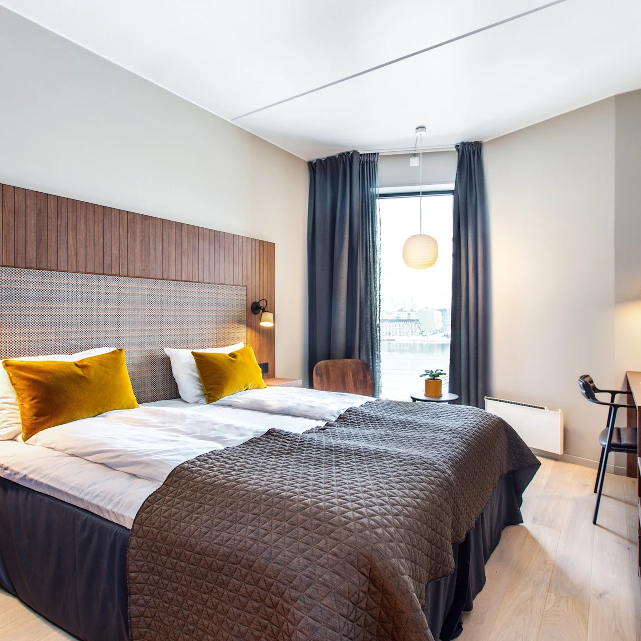 Et pent arrangert hotellrom med dobbeltseng med gule puter, et trepanel, sengebordlamper, et skrivebord med lampe og et stort vindu med gardiner.