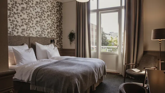 Et komfortabelt hotellrom med en stor seng med hvitt sengetøy, blomstret tapet og et lyst vindu med gardiner.