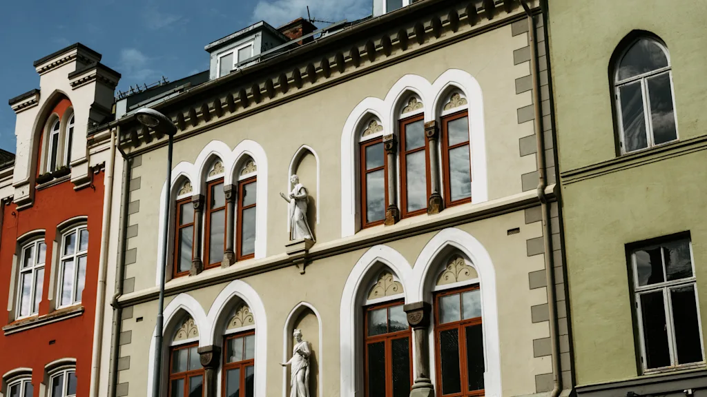 Udsmykkede bygningsfacader med buede vinduer og dekorative statuer under en blå himmel. En blanding af arkitektoniske stilarter og farver.