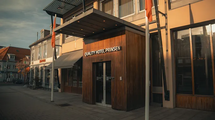 Hotellin sisäänkäynti, jossa lukee "QUALITY HOTEL PRINSEN" puuverhoilulla, sijaitsee rauhallisella kadulla, jossa on toinen rakennus merkinnällä "NAPOLI" lähistöllä.