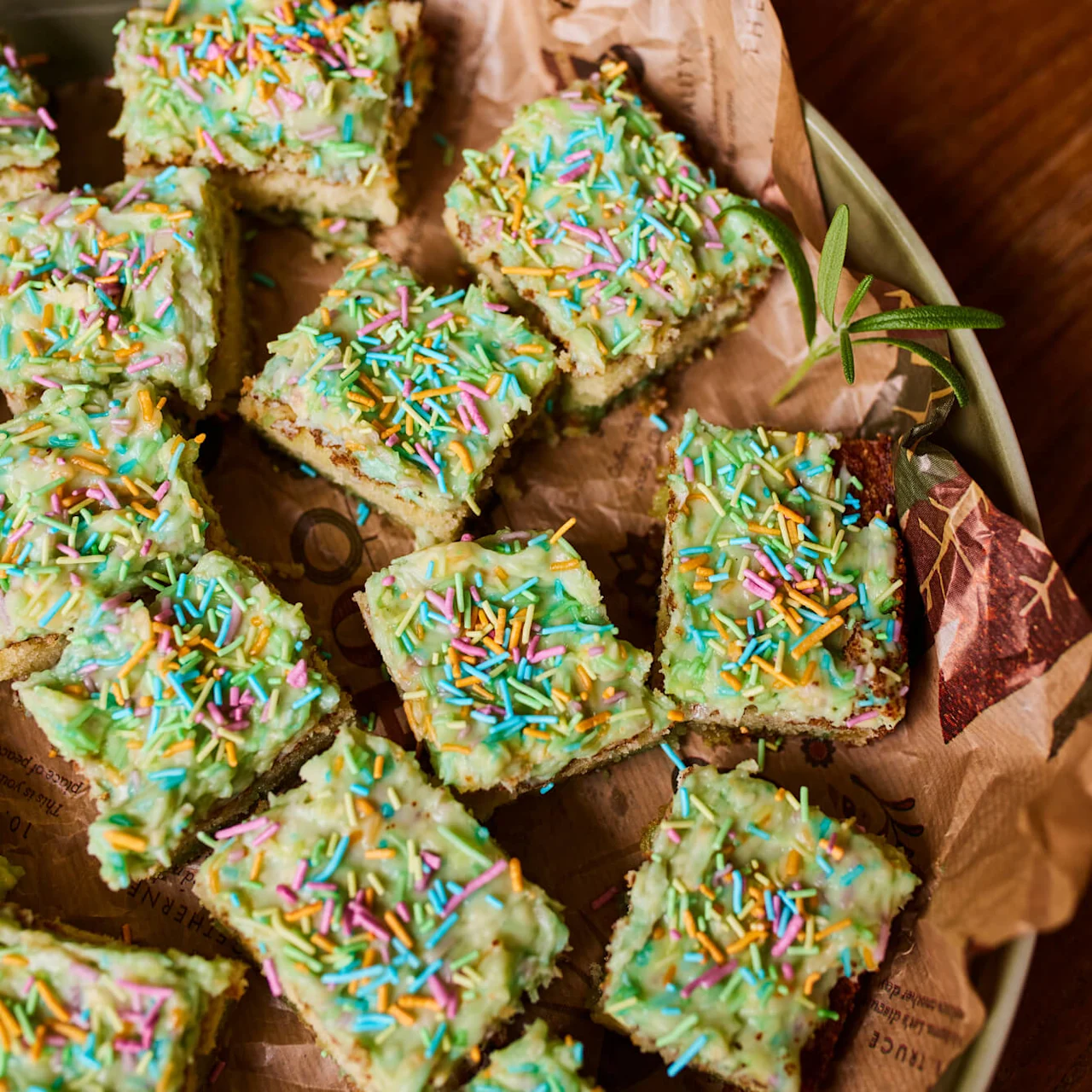 Fargerike kakeruter med frosting og regnbuestrøssel ligger på dekorert papir i et rundt brett. En grønn plante i nærheten gir en naturlig touch til en treflate.