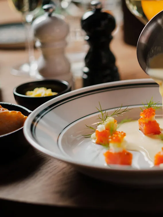 A person pours sauce from a copper pan onto a dish with garnished salmon cubes on a table set with salt, pepper shakers, bread, and glassware.