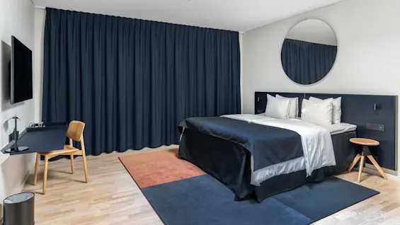 Et moderne hotelværelse med en stor seng, mørkeblå gardiner, et rundt spejl og et skrivebordsområde.