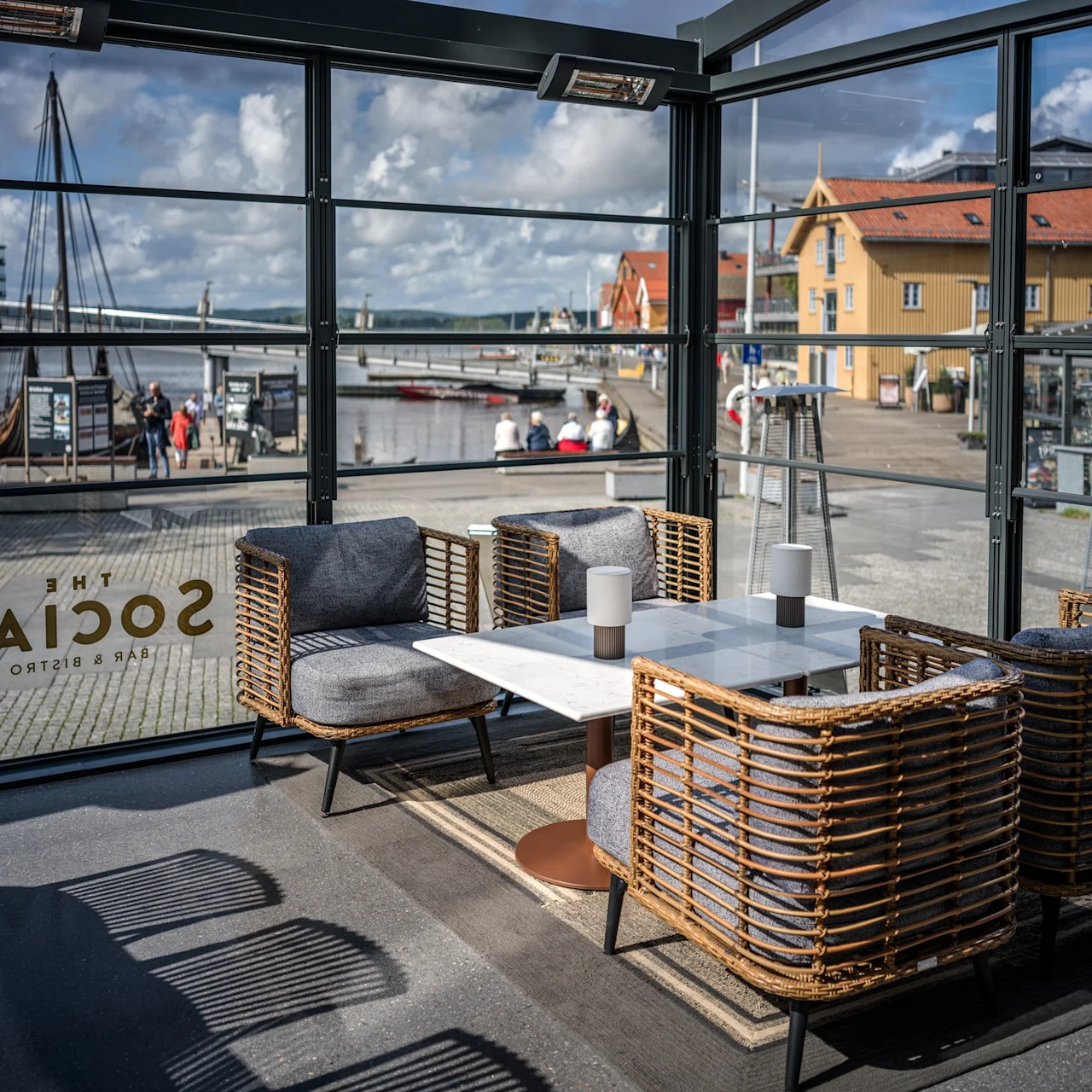 Korgstolar omger ett marmorbord i ett glasomslutet område märkt "THE SOCIAL BAR & BISTRO," med utsikt över en flodmiljö med en bro, förtöjda båtar och färgglada byggnader.