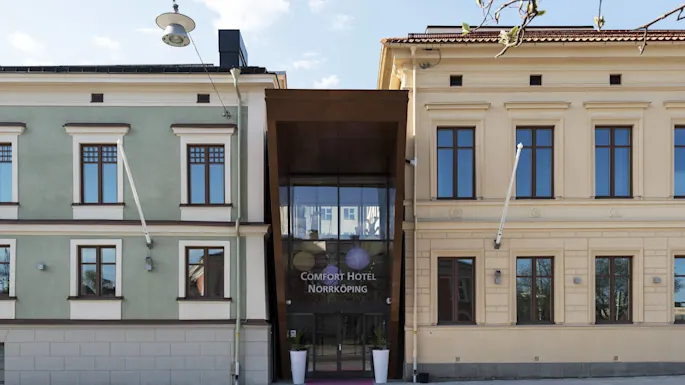 Inngang til Comfort Hotel med en rosa løper og to plantekasser, i en urban setting med historiske bygninger.