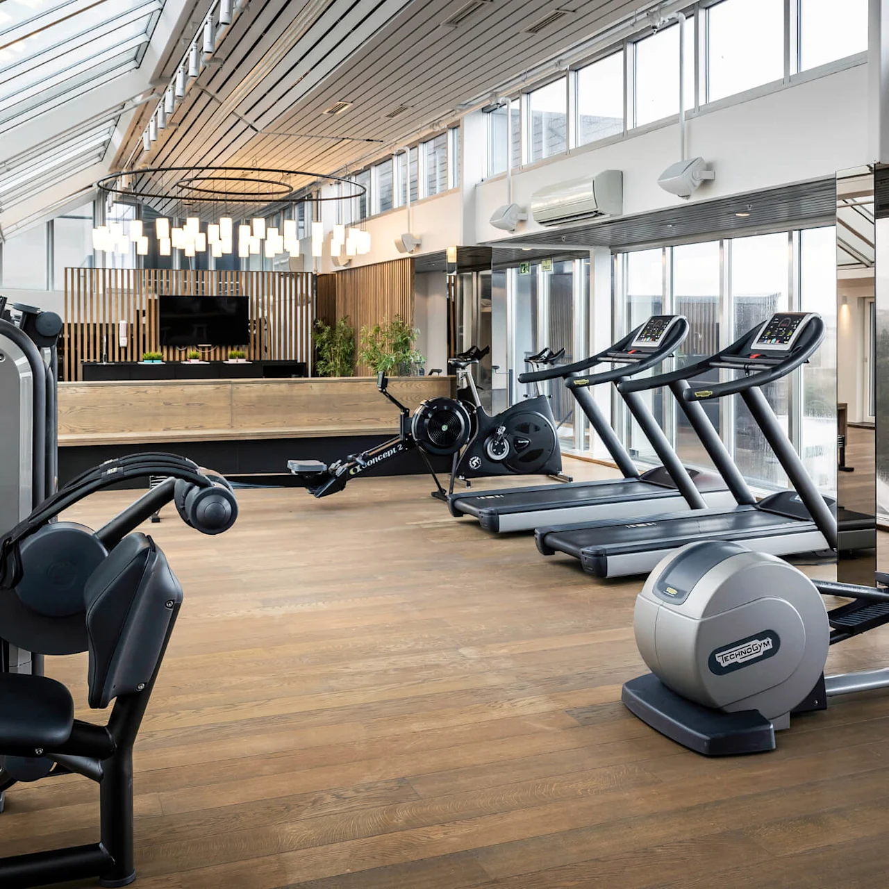 Tredemøller, ellipsemaskin og forskjellige treningsutstyr er plassert på et tregulv i et solrikt rom med store vinduer og taklamper. "Technogym"-merking er synlig på flere maskiner.