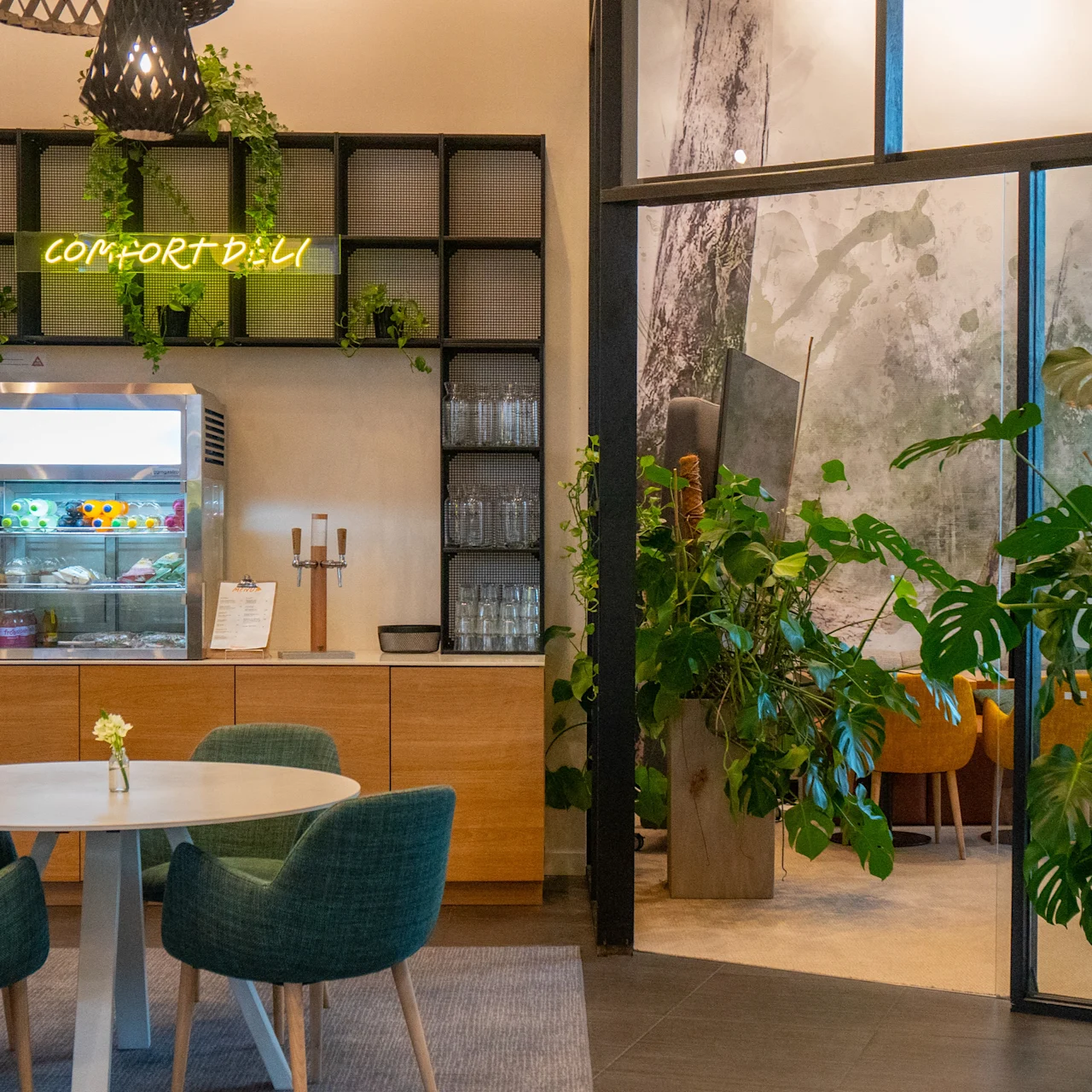 A café area features a refrigerated display with snacks and drinks labeled "Comfort Deli." Shelves hold glasses and plants. Green chairs surround a round table. Nearby, large plants enhance the ambient aesthetic. 