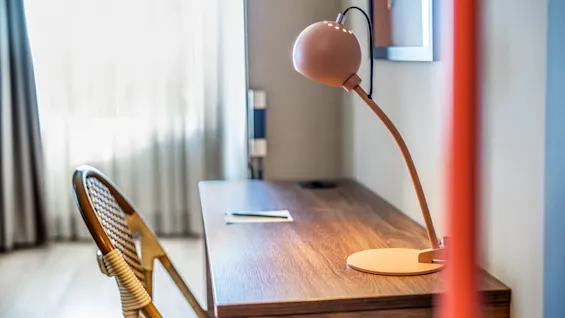 A desk with a curved lamp and a chair stands in a well-lit room with thin curtains covering a window.