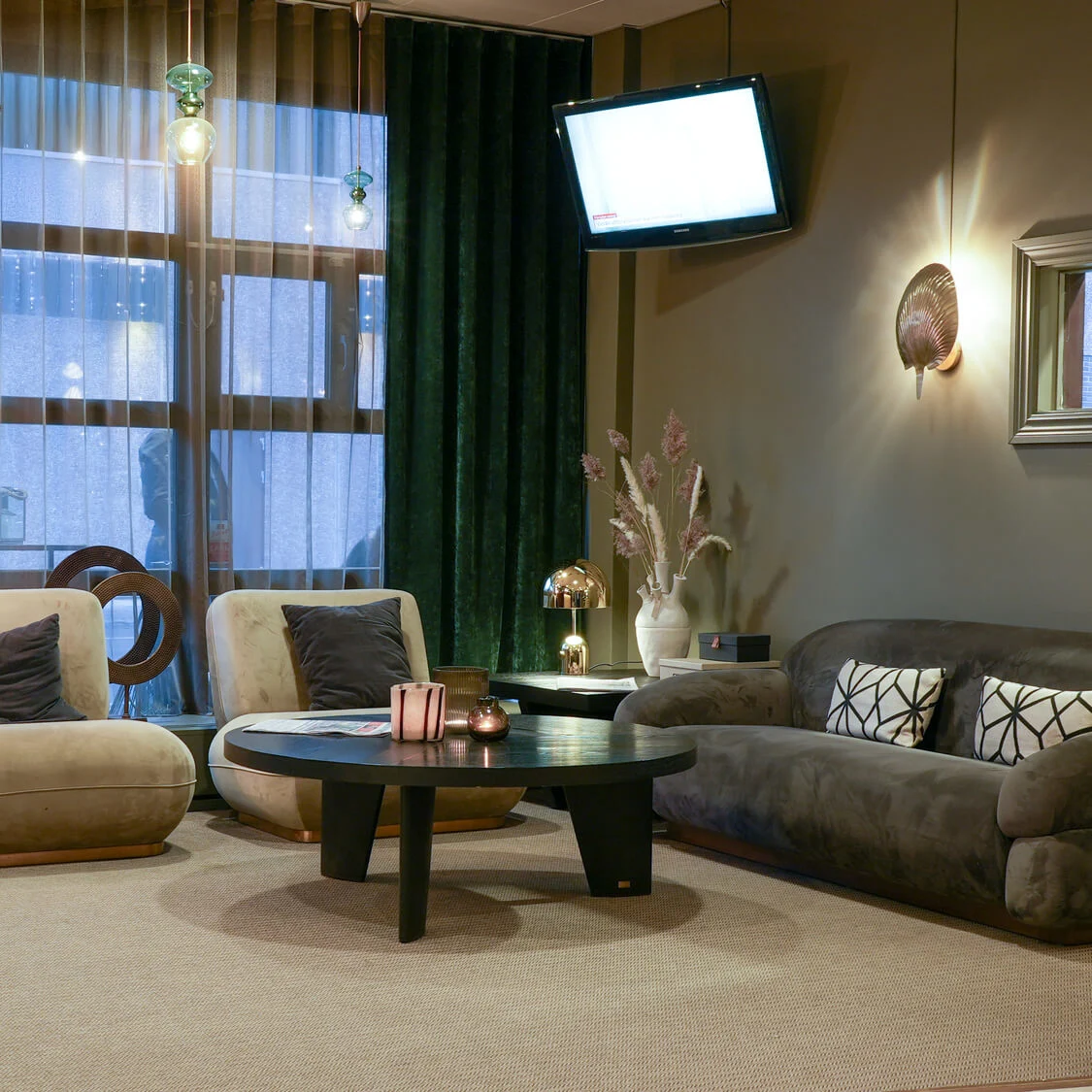 En hyggelig stue med beige stole, en mørk sofa med mønstrede puder og et sort sofabord. Overhead-belysning og dekorative elementer tilføjer varme mod et stort vinduesbaggrund.