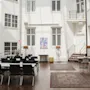 En solbelyst atrium har rektangulära bord med svarta stolar, omgivna av vita väggar med flera fönster. Mattor täcker trägolvet, och konstverk hänger längs bakväggen.