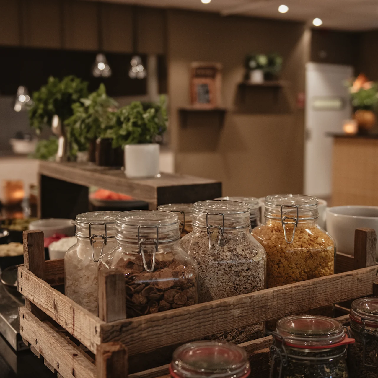 Glass jars filled with various grains and cereals sit in a wooden crate on a kitchen counter, surrounded by herbs and modern kitchen decor.