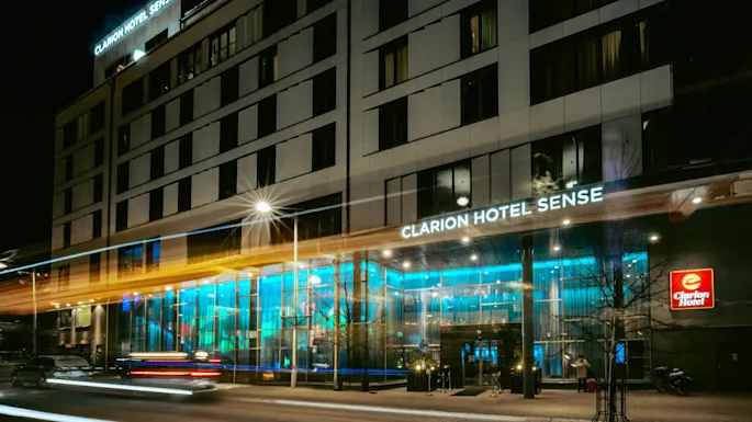 Eksteriøret til et opplyst hotell med skiltet "CLARION HOTEL SENSE". Biler med lysstriper kjører forbi på en snørik natt. Et rødt Clarion Hotel-skilt er synlig.