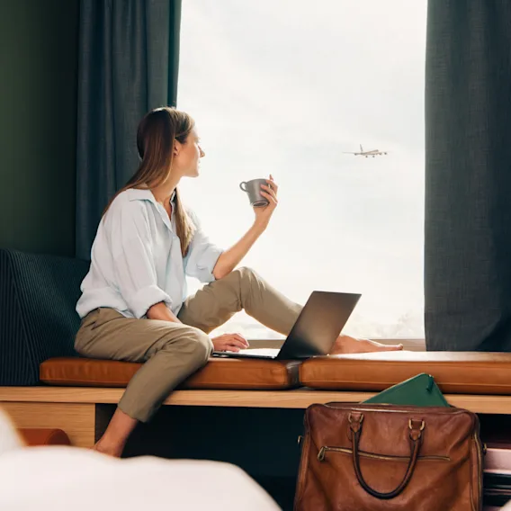 Person som jobber på en bærbar datamaskin ved et vindu, holder en kopp og ser et fly fly forbi.