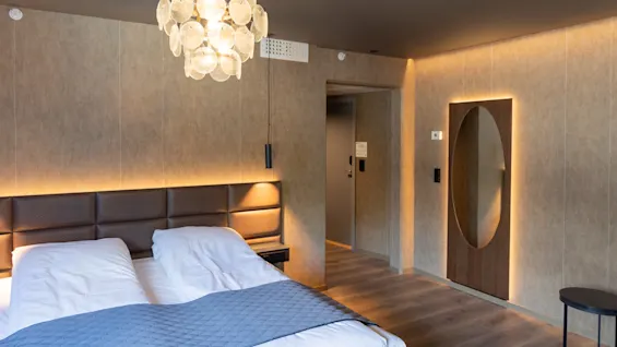 En pent dobbeltseng med quiltet hodegjerde står under en moderne lysekrone, i et varmt belyst rom med tregulv, ovalformet speil og minimalistiske møbler.