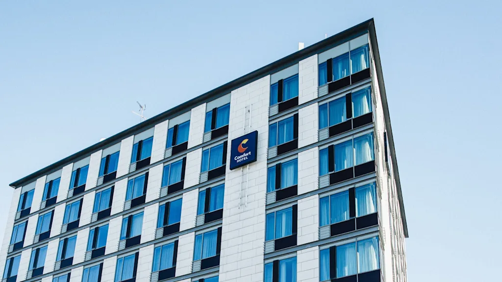 A multistory building stands with a sign reading "Comfort Hotel," featuring blue-tinted windows, set against a clear sky.