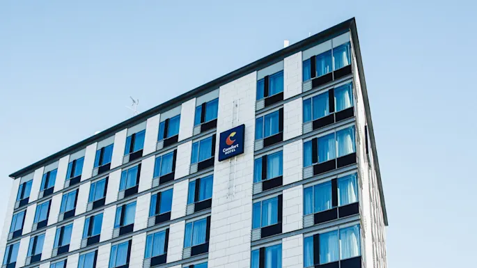 Useamman kerroksen rakennus, jossa on kyltti "Comfort Hotel", sinertävillä ikkunoilla, kirkasta taivasta vasten.