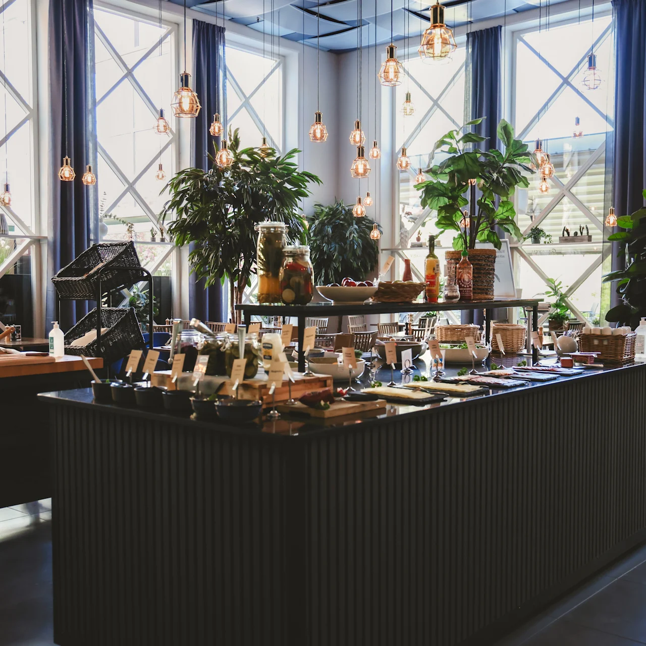 A buffet table displays various dishes under warm lighting in a bright room with large windows and potted plants. Hanging lights add ambiance.
