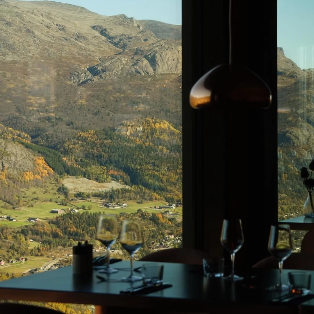 Dining tables with glassware rest against large windows, framing a vast mountain landscape with lush greenery and rocky peaks under a clear blue sky.