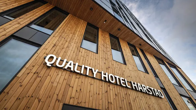 A wooden building facade with large windows displays the sign "QUALITY HOTEL HARSTAD" against a cloudy sky backdrop.