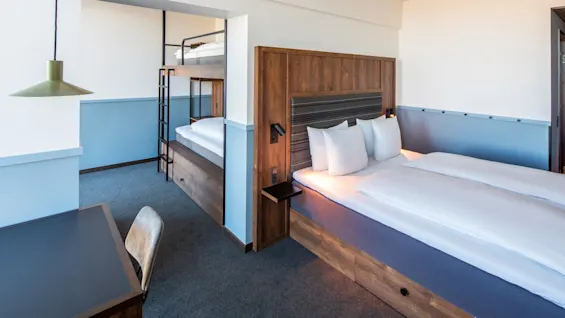 Dobbeltseng med hvitt sengetøy står på et hotellrom ved siden av en køyeseng i tre; et skrivebord med stol og en grønn hengelampe kompletterer den moderne innredningen.