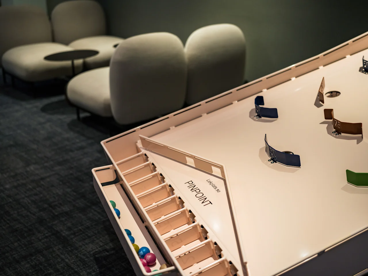 Et brettspill med teksten "Miniature PinPoint" i et svakt opplyst rom, med fargerike små baller og kurvete baner. Bakgrunnen har myke sitteplasser og små bord.