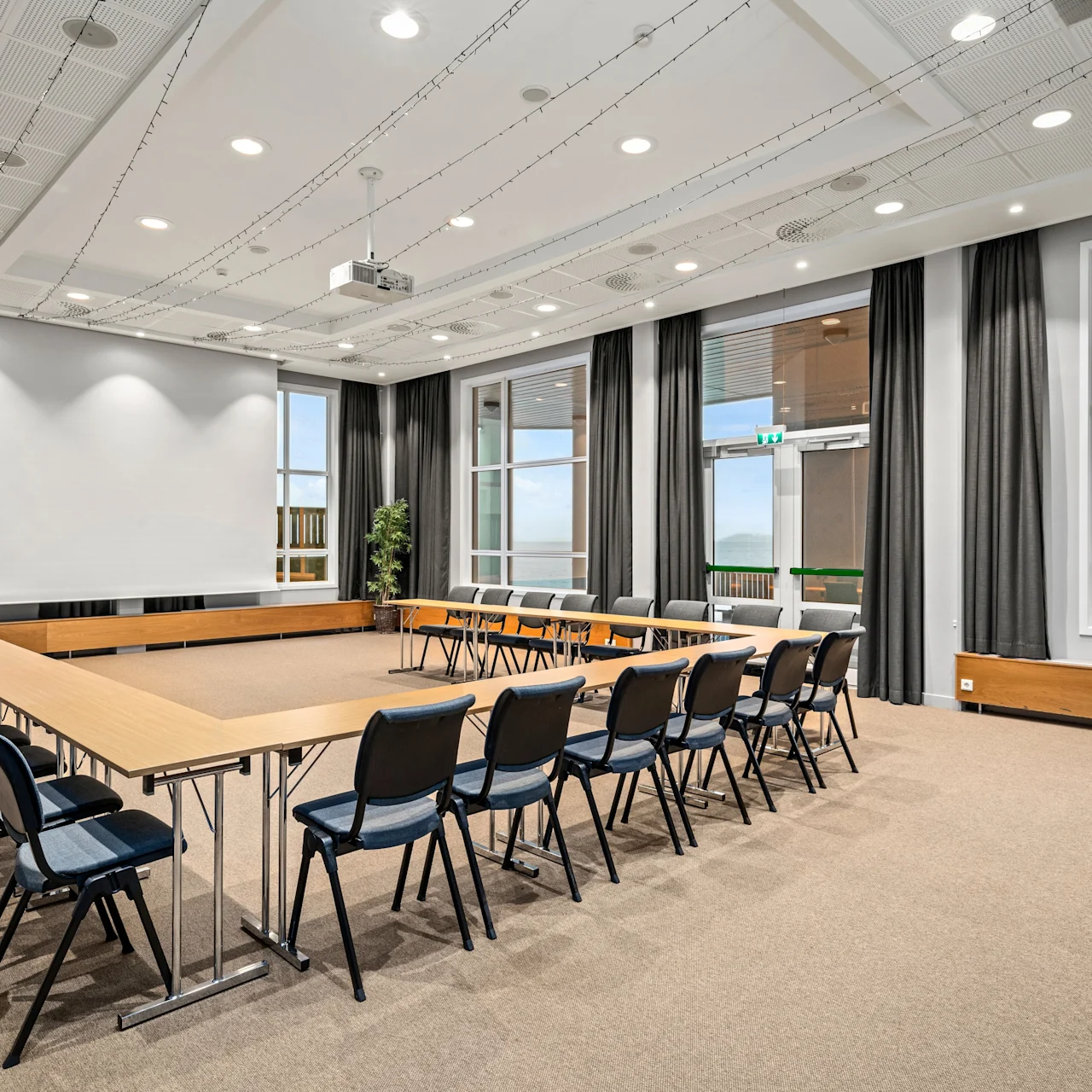 Konferencebord omgivet af stole i U-form; store vinduer med gardiner afslører en lys udsigt. Loftslamper og en projektor forbedrer det moderne mødelokale.