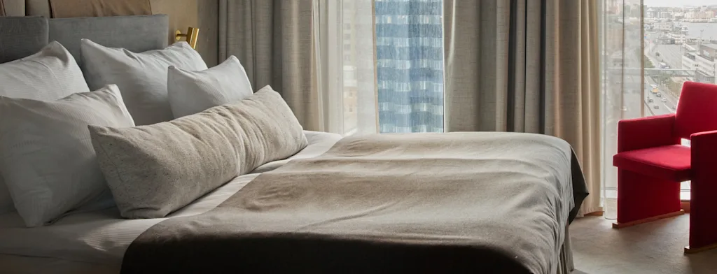 Moderni hotellihuone, jossa on mukava sänky, eloisa punainen nojatuoli ja suuret ikkunat kaupunkinäköalalla.