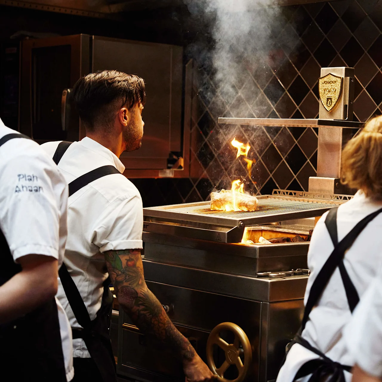 Kokke i et køkken betragter en flammende grill med fokuseret opmærksomhed; et travlt, professionelt kokkemiljø.