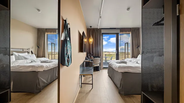 Et moderne hotellrom med to senger med hvite laken vendt mot et stort vindu som viser en naturskjønn utsikt. Et skrivebord med stol og en hengende lampe ved siden av. Refleksjoner i et speil.